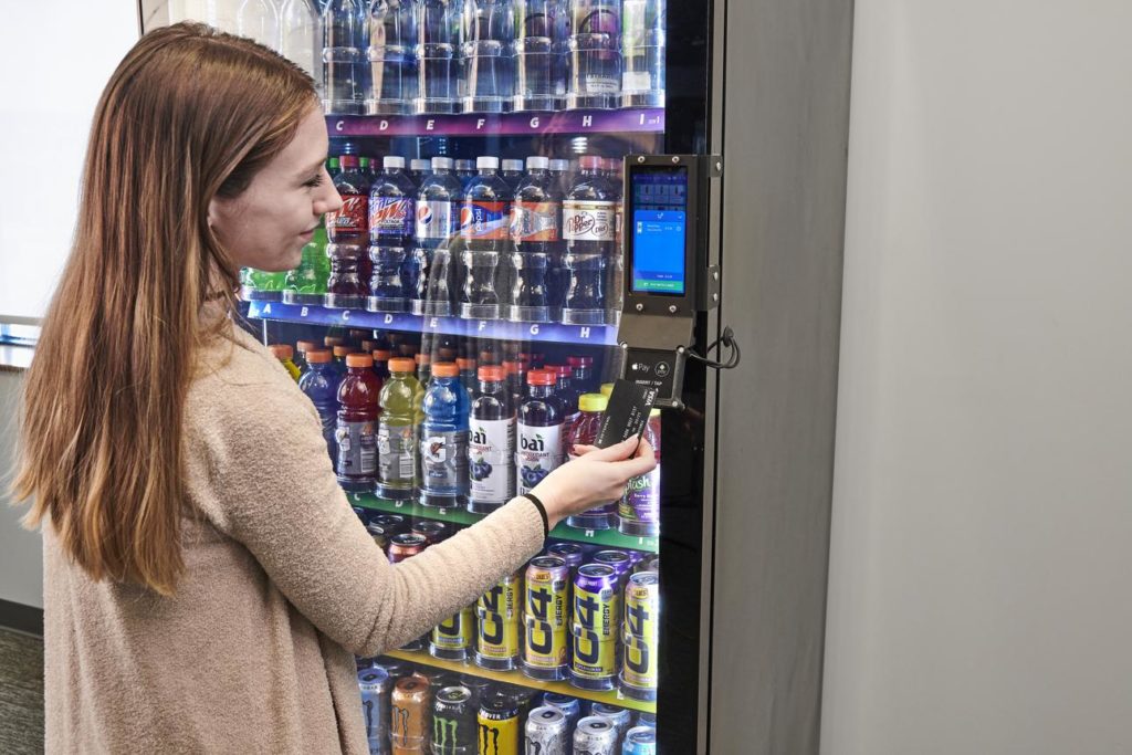 Employee making a contactless payment at a modern drinks vending machine in a commercial workplace.